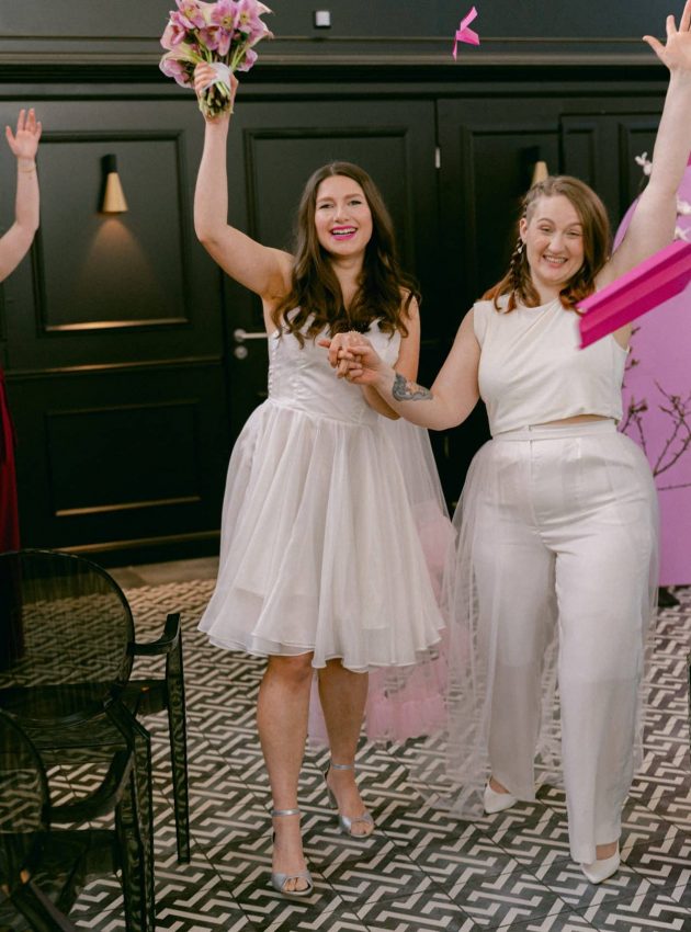 zwei Frauen beim Auszug der freien Trauung, sie tragen weiße Hochzeits-Outfits und werden von Papierfliegern begleitet, die ihre Gäst*innen werfen