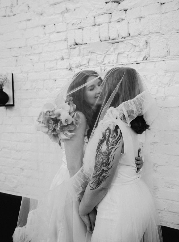 zwei weiblich gelesene Personen sind im Begriff, sich zu küssen. Sie lehnen unter einem Schleier an einer groben, geweißten Backsteinwand.