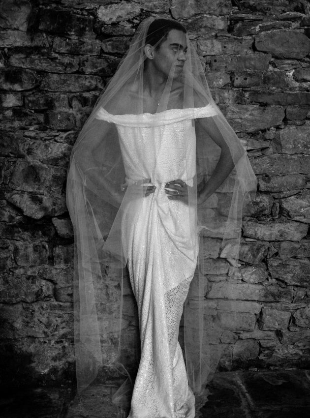 Männlich gelesene Schwarze Person lehnt an einer groben Bruchsteinwand. Sie trägt ein schulterfreies, bodenlanges Paillettenkleid und einen langen Schleier. Die Arme in die Hüften gestemmt, den Kopf zur Seite gedreht.