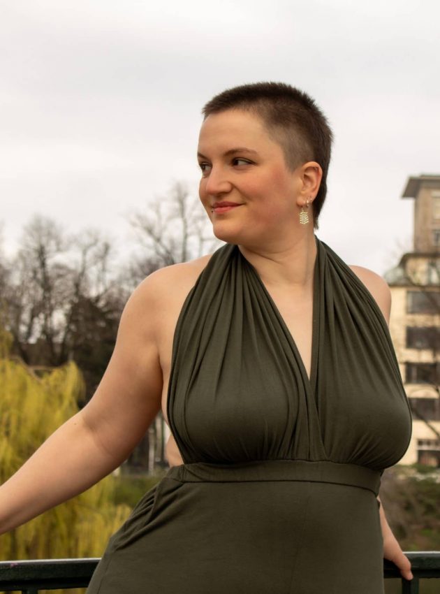 Christina trägt einen dunkelgrünen Infinity-Jumpsuit als Neckholder. Sie lehnt am Geländer einer Brücke im Park.